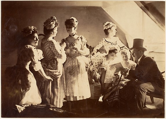 [A Group of Six Costumed Women Posed in Interior with Top Hatted Gentlemen]