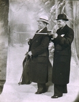 [Two Men in Overcoats, One Gesturing with Cane, Posed in Front of Painted Backdrop]
