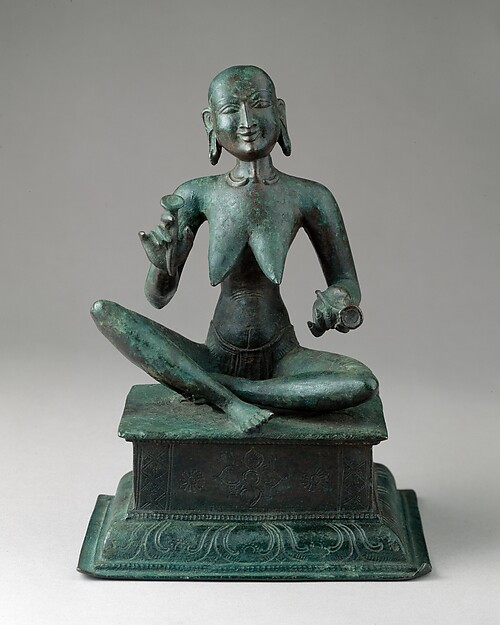 A small, extremely thin woman sits with one leg raised and a pair of cymbals in her hands. Her ribs and joints are sharply defined, while her face turns upward with an animated smile.