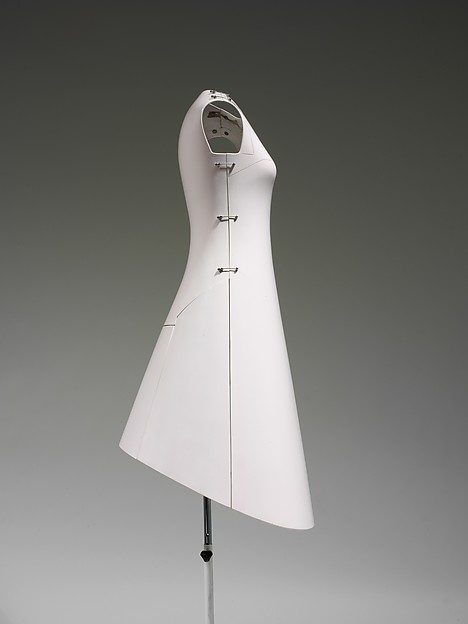 Side profile of the closed dress on a stand, a smooth hard shell flaring from a fitted bodice to an angled hem, with metal clasps along a vertical seam.
