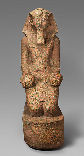 A monumental granite figure of Hatshepsut kneels upright and holds rounded offering vessels. She wears the striped royal headcloth and ceremonial false beard associated with an Egyptian king.
