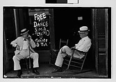 [One 35mm Film Frame: Two Elderly Men in Conversation on Porch in Front of Painted Sign for 