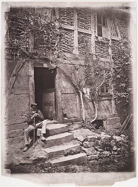 [Man Sitting on Steps of House with Socks Hanging on Nearby Vine to Dry]
