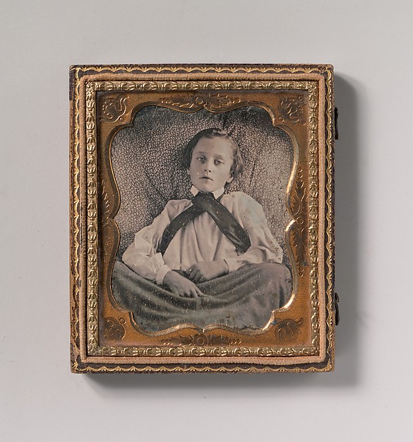 [Boy Seated Cross-legged, Partially Covered by Blanket, Leaning Against Cushion]