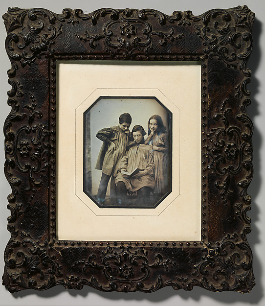 [Three Children, One Seated Holding an Open Book, the Other Two Standing, in Front of a Light Background]