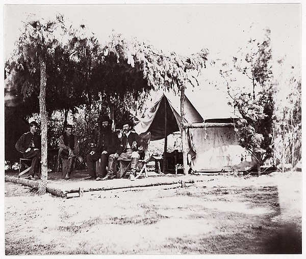 [Four men in camp under a lean-to of pine boughs]